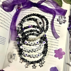 Puede incluir: Una colección de pulseras de cuentas en negro, transparente y morado. Algunas pulseras deletrean "BTS" y "JUNGKOOK" con cuentas de letras blancas. Los dijes de estrella y las cuentas en forma de estrella agregan detalles. Las pulseras están dispuestas sobre un libro abierto con una cinta morada.
