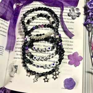 Puede incluir: Conjunto de pulseras de cuentas negras, blancas y moradas. Algunas pulseras deletrean "BTS" y "JHOPE" con cuentas de letras. Los dijes de estrella y las cuentas añaden detalles. Las pulseras están sobre un libro abierto con cinta morada y flores.