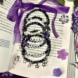 Puede incluir: Una colección de pulseras de cuentas negras, blancas y moradas. Algunas pulseras tienen colgantes de estrella y cuentas de letras que deletrean "BTS", "RM" y "BNGTON". Las pulseras están dispuestas sobre un libro abierto con cinta morada y detalles florales.