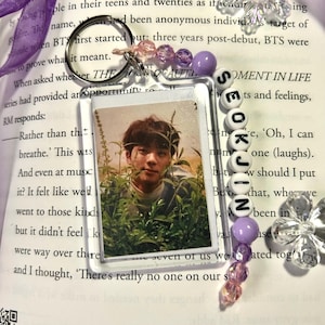 May include: A clear acrylic keychain featuring a photo of a person surrounded by greenery. The keychain includes a silver keyring, purple and pink beads, and white letter beads spelling "SEOKJIN". The background is a book.