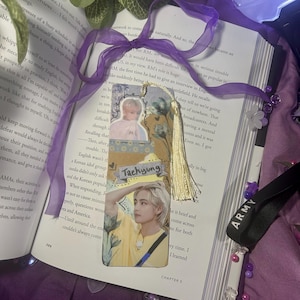 May include: A handmade bookmark featuring two images of a person, with the name "Taehyung" printed on it. The bookmark has a gold tassel and is decorated with a purple ribbon. The bookmark is placed on an open book.