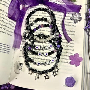 Puede incluir: Varios brazaletes de cuentas negras, transparentes y moradas, con dijes de estrella y corazón, se exhiben sobre un libro abierto. Algunos brazaletes deletrean "BTS" y "V". Una cinta morada y flores decorativas complementan la escena.
