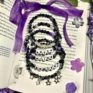 Puede incluir: Varios brazaletes de cuentas negras, blancas y moradas están colocados sobre un libro abierto. Algunos brazaletes deletrean "BTS" y "JIN" con acentos de corazón. Los dijes de estrella plateada y una cinta morada completan el diseño.