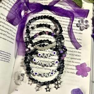 Puede incluir: Una colección de pulseras de cuentas con cuentas negras, transparentes y moradas. Algunas pulseras presentan cuentas de letras blancas que deletrean "BTS", "JIMIN" y "BONGTON". Encantos de estrellas plateadas cuelgan de las pulseras. Las pulseras están sobre un libro abierto.