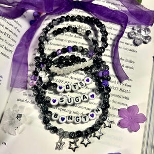 Puede incluir: Una colección de pulseras de cuentas en negro, transparente y morado. Algunas pulseras tienen cuentas de letras blancas que deletrean "BTS", "SUGA" y "BONGT". Dijes de estrella plateada cuelgan de una pulsera. Las pulseras están sobre un libro abierto.