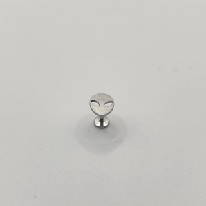 May include: A silver-colored stud earring featuring an alien head design. The earring has a flat, circular base and a post for insertion. The alien head has large, oval eyes and a small, curved mouth. The earring is likely made of stainless steel.
