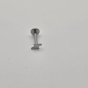 May include: A silver-colored lip piercing stud with a flat top and a lightning bolt design on the bottom. The jewelry is designed for body modification and is shown against a white background.