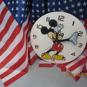 May include: A round wall clock featuring Mickey Mouse in a classic pose, with black hands and a red and white outfit. The clock face has black numerals. The background includes American flags with red and white stripes and blue fields of stars.