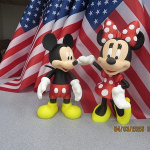 Peut inclure: Figurines de Mickey et Minnie Mouse devant des drapeaux américains. Mickey est en noir, rouge, jaune et blanc. Minnie porte une robe à pois rouge, un nœud rouge et des chaussures jaunes. La date 04/03/2026 08:02 est visible.