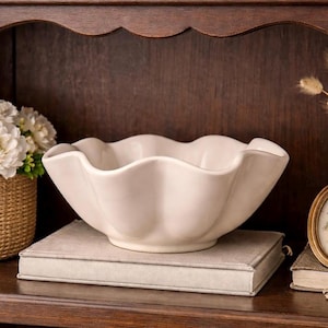 Peut inclure: Un bol en céramique crème avec un bord festonné. Le bol est posé sur un livre de couleur claire. L'arrière-plan est une armoire en bois foncé. Un petit panier de fleurs blanches se trouve à gauche du bol.