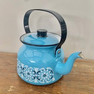 May include: A light blue enamel teapot with a black handle and lid. The teapot has a decorative white and blue floral pattern around the base. The spout has a gold-coloured trim. The teapot is on a wooden surface.