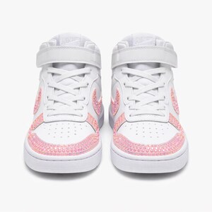May include: Pair of white high-top sneakers with pink rhinestone accents. The sneakers feature a strap closure and the Nike logo on the tongue. The toe and side panels are embellished with pink rhinestones.