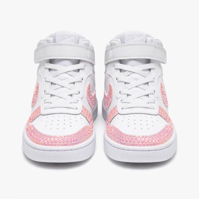 May include: Pair of white high-top sneakers with pink rhinestone accents. The sneakers feature a strap closure and the Nike logo on the tongue. The toe and side panels are embellished with pink rhinestones.