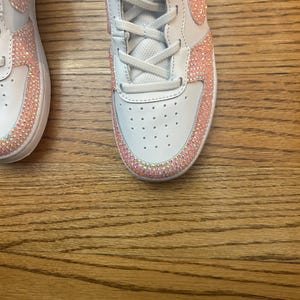 May include: White sneakers with pink rhinestone accents. The shoes have white laces and a pink Nike swoosh. The rhinestones are along the edges of the shoe and around the swoosh. The shoes are on a wooden surface.
