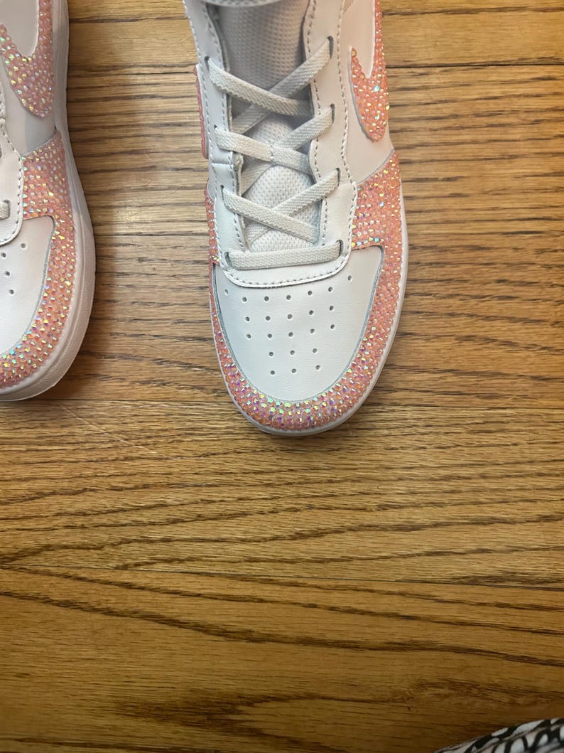 May include: White sneakers with pink rhinestone accents. The shoes have white laces and a pink Nike swoosh. The rhinestones are along the edges of the shoe and around the swoosh. The shoes are on a wooden surface.