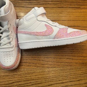 May include: White high-top sneakers with pink rhinestone accents. The Nike swoosh and the toe box are embellished with sparkling pink rhinestones. The sneakers have a white sole and a velcro strap.