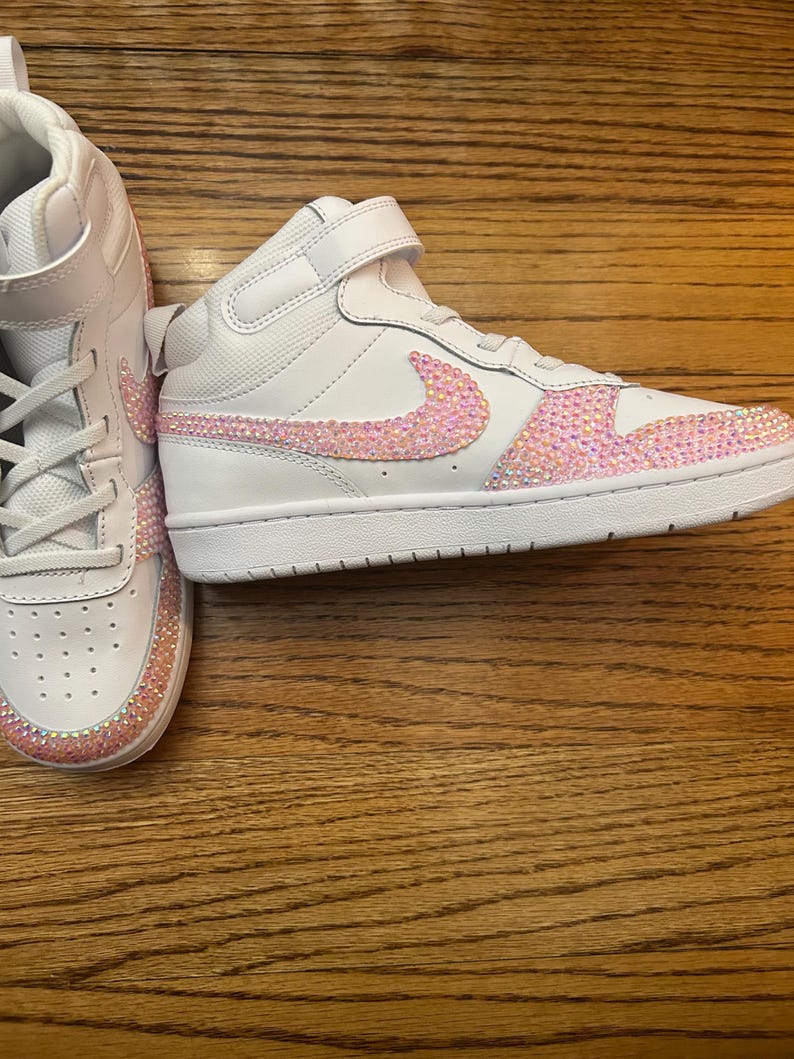 May include: White high-top sneakers with pink rhinestone accents. The Nike swoosh and the toe box are embellished with sparkling pink rhinestones. The sneakers have a white sole and a velcro strap.