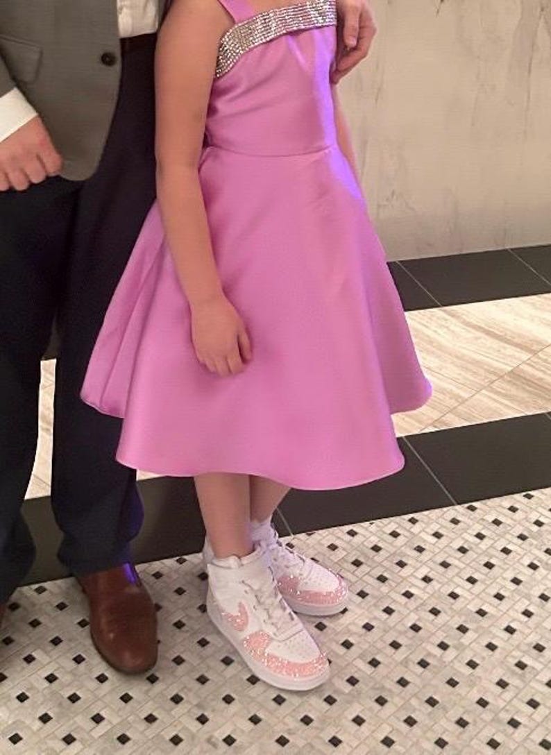 May include: A light pink dress with a sparkling strap and a flared skirt. The dress is paired with white sneakers adorned with pink embellishments. The image captures a formal or celebratory occasion.