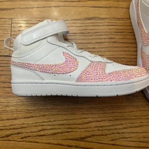 May include: White high-top sneakers with a pink crystal-like design on the Nike swoosh and toe area. The sneakers have a velcro strap and the Nike logo on the tongue.