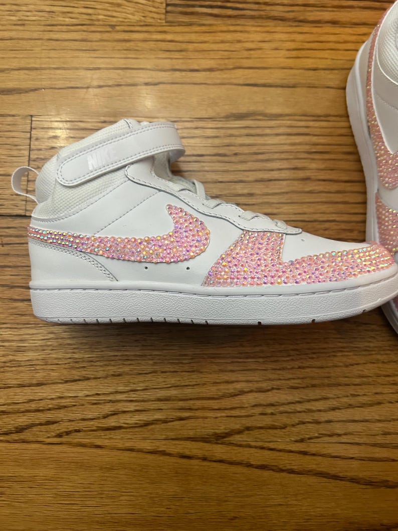 May include: White high-top sneakers with a pink crystal-like design on the Nike swoosh and toe area. The sneakers have a velcro strap and the Nike logo on the tongue.