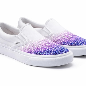 May include: White slip-on sneakers with a gradient of purple and blue rhinestones on the toe. The shoes have a white sole and the Vans logo is visible on the side. A stylish and eye-catching footwear choice.