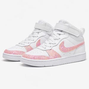 May include: White high-top sneakers with pink rhinestone accents on the Nike swoosh and around the soles. The sneakers have a velcro strap and white laces. The shoes are set against a white background.