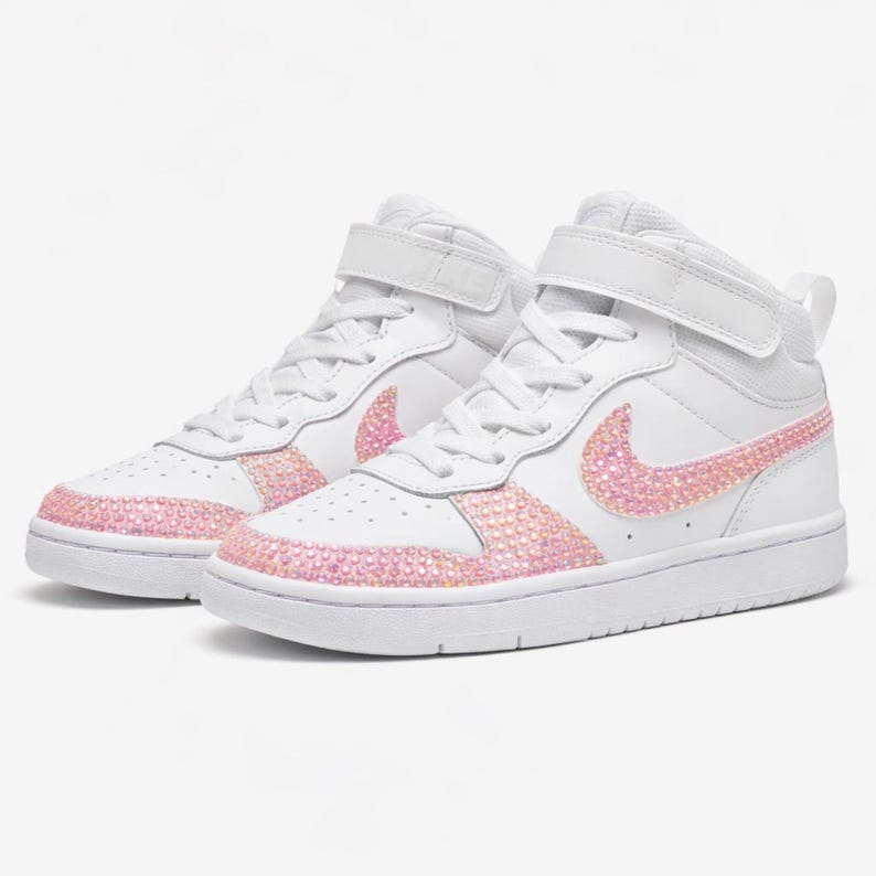 May include: White high-top sneakers with pink rhinestone accents on the Nike swoosh and around the soles. The sneakers have a velcro strap and white laces. The shoes are set against a white background.