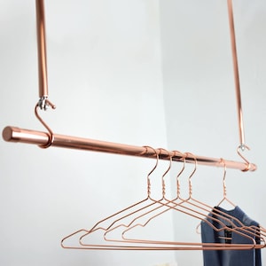 May include: A copper-colored clothing rack suspended from the ceiling. The rack has a horizontal bar with several matching wire hangers. A navy blue garment hangs on the hangers. The rack is against a white wall.