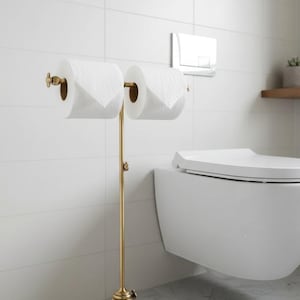 May include: A gold-colored, free-standing toilet paper holder with two rolls of white toilet paper. The holder has a decorative base and a vertical pole with curved arms to hold the rolls. The background is a white tiled wall and a white toilet.