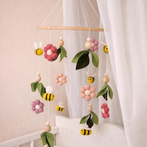 May include: A handmade baby mobile featuring felt flowers, green leaves, and yellow bees. The mobile is suspended from a wooden bar with white strings. The flowers are pink and white, creating a whimsical nursery decoration.
