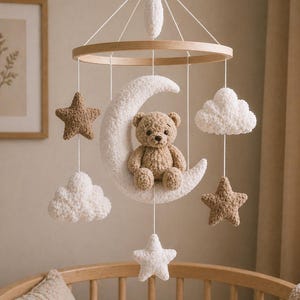 Puede incluir: Móvil para bebé hecho a mano con un oso de peluche sobre una luna creciente, estrellas y nubes. El móvil está suspendido de un anillo de madera y presenta colores suaves y neutros.