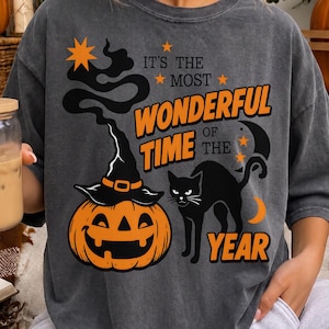 May include: A charcoal gray t-shirt featuring a graphic print of a jack-o'-lantern wearing a witch hat, a black cat, and the text "IT'S THE MOST WONDERFUL TIME OF THE YEAR" in orange and black, accented with stars and a crescent moon.