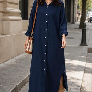 May include: A long-sleeved navy blue button-down shirt dress featuring a classic collar, a full-length front button placket with white buttons, and side slits at the hem. The garment is worn with a brown and cream shoulder bag and beige mules.