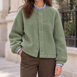 May include: A sage green fleece jacket with a round neckline and long sleeves. The jacket features a front button closure with five flower-shaped buttons. Underneath, a blue and white striped collared shirt is visible at the neck and cuffs.