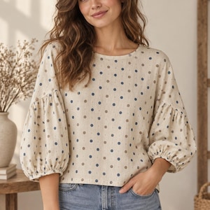 May include: A beige long-sleeve blouse featuring a repeating pattern of small blue and brown polka dots. The garment has a round neckline and voluminous, gathered puff sleeves that end in elasticated cuffs, creating a relaxed, loose-fitting silhouette.
