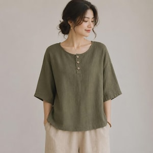 May include: An olive green short-sleeved blouse featuring a round neckline and a partial front placket with three small, light-coloured buttons. The garment has a relaxed fit with elbow-length sleeves and is paired with light beige trousers.