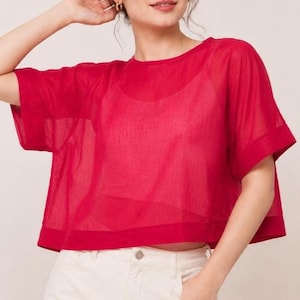 May include: A vibrant red short-sleeved top featuring a sheer, lightweight fabric with a loose, boxy fit and a rounded neckline. The garment is layered over a matching red base layer and is paired with light-colored pants with a visible button.