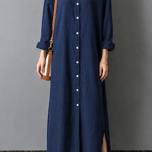 May include: A long-sleeved navy blue button-down shirt dress featuring a classic collar, a full-length front button placket with white buttons, and side slits at the hem. The garment is paired with a brown leather shoulder bag and beige mules.
