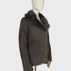 May include: A dark brown suede jacket featuring a wide, plush dark gray faux fur collar and matching fur trim at the cuffs and hem. The jacket includes an asymmetrical front zipper closure and long sleeves, displayed on a mannequin form.