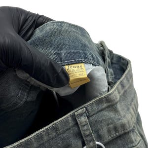 May include: Close-up of a pair of faded blue jeans. The jeans have a light wash and visible stitching. A yellow tag with the word "Jeans" is visible. The jeans are held by a gloved hand.