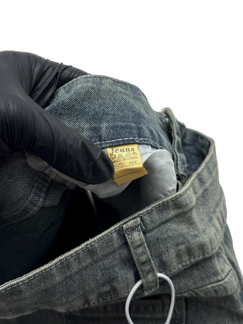May include: Close-up of a pair of faded blue jeans. The jeans have a light wash and visible stitching. A yellow tag with the word "Jeans" is visible. The jeans are held by a gloved hand.