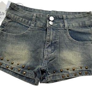 May include: Faded denim shorts with a button closure and front pockets. The shorts are embellished with bronze-colored studs along the bottom hem. The shorts have a distressed look.