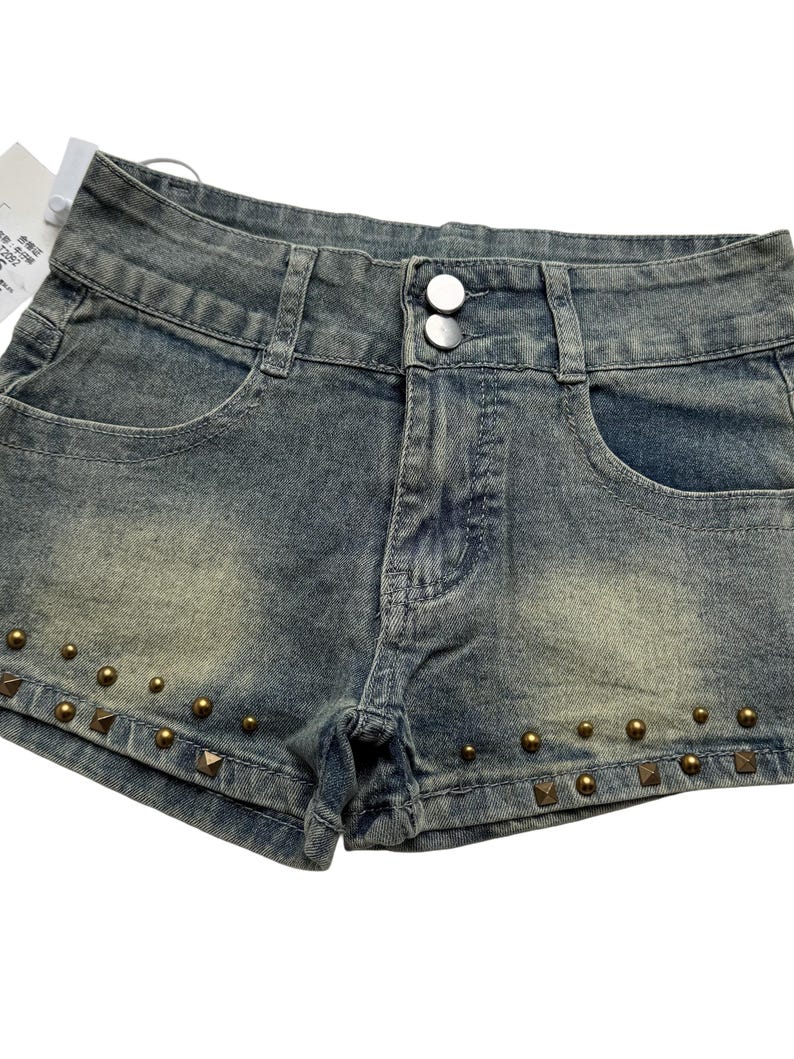May include: Faded denim shorts with a button closure and front pockets. The shorts are embellished with bronze-colored studs along the bottom hem. The shorts have a distressed look.