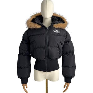 Peut inclure: Veste matelassée noire avec capuche bordée de fausse fourrure. La veste courte présente un ourlet et des poignets côtelés. Le mot "SRIKER" est brodé en blanc sur la poitrine. La veste est présentée sur un mannequin.
