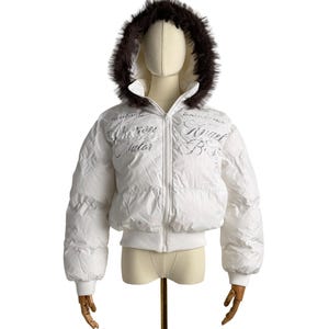 May include: White puffer jacket with a faux fur-trimmed hood. The jacket features a zip-up front, ribbed cuffs, and the words "Merone Salar" and "Angel Boss" printed on the front. The jacket is displayed on a mannequin.