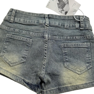 May include: Close-up of denim shorts with a faded wash. The shorts have two back pockets and a waistband with belt loops. A white tag is attached to the shorts.
