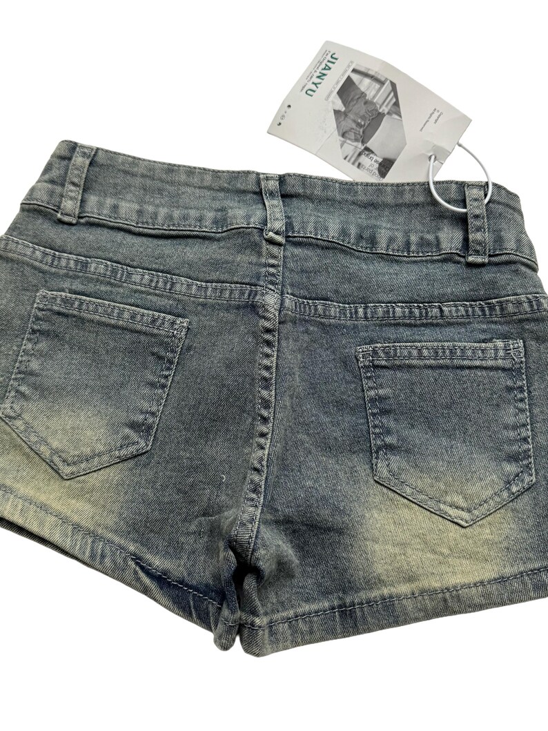 May include: Close-up of denim shorts with a faded wash. The shorts have two back pockets and a waistband with belt loops. A white tag is attached to the shorts.