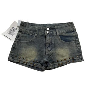 May include: Faded denim shorts with a button closure and front pockets. The shorts are embellished with small, bronze-colored studs along the bottom hem. A white tag is attached to the shorts.