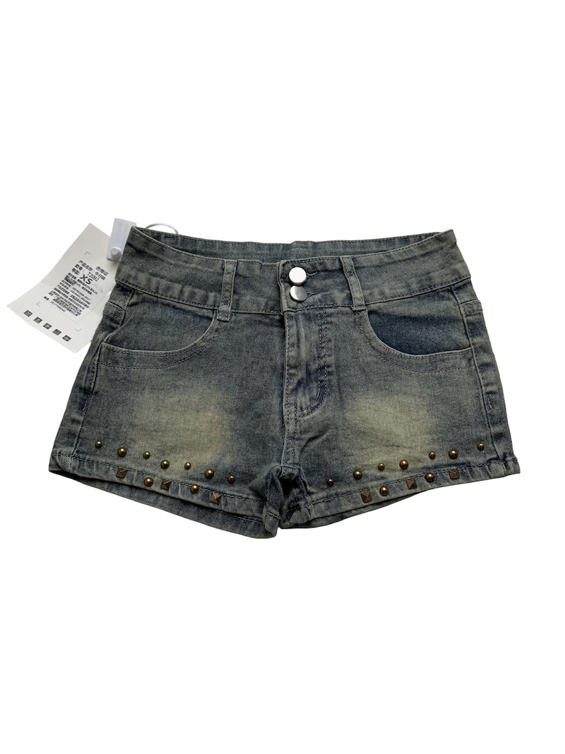 May include: Faded denim shorts with a button closure and front pockets. The shorts are embellished with small, bronze-colored studs along the bottom hem. A white tag is attached to the shorts.