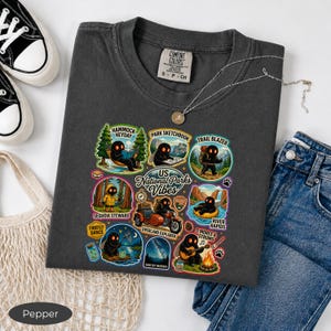May include: A charcoal gray crew neck t-shirt featuring a graphic print titled US National Parks Vibes. The design includes multiple circular illustrations of a black creature engaging in outdoor activities like hiking, camping, and stargazing.